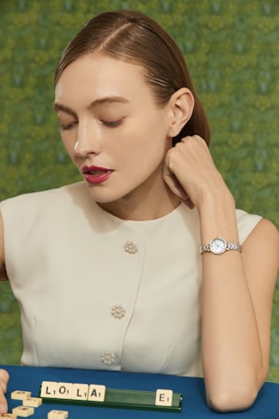 A young woman with short, slicked-back hair is dressed in a sleeveless, white top adorned with pearl-like buttons. She rests her chin on her hand, which bears an elegant wristwatch. In front of her on a blue surface lies a row of letter tiles spelling out 'LOLA', along with an additional tile featuring the letter 'E'. The backdrop is a blurred, textured green pattern.