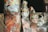 Several colorful traditional puppets with elaborate headdresses wrapped in plastic are arranged in close proximity. The setting appears to be a storage or market area with some parts of the background slightly blurred.