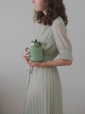 A woman with curly hair is seen in profile, wearing a light green pleated dress. She is holding a small green potted plant close to her body. The background is minimalistic and plain, enhancing the serene and calming atmosphere.