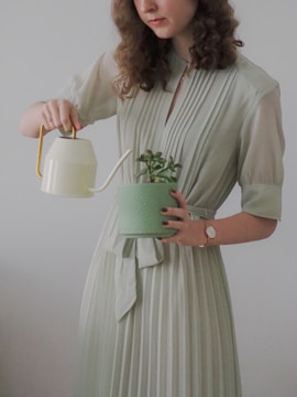 A person with curly hair wearing a light green dress is watering a small plant in a textured green pot using a light yellow watering can. Their other hand wears a wristwatch with a brown strap.