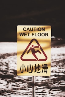 A caution sign warning of a wet floor stands prominently. The sign is yellow with a red graphic showing a figure slipping. English text 'Caution Wet Floor' is written above, and Chinese text is displayed below the graphic. The background is a blurred image of a watery surface.