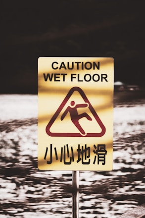 A caution sign warning of a wet floor stands prominently. The sign is yellow with a red graphic showing a figure slipping. English text 'Caution Wet Floor' is written above, and Chinese text is displayed below the graphic. The background is a blurred image of a watery surface.