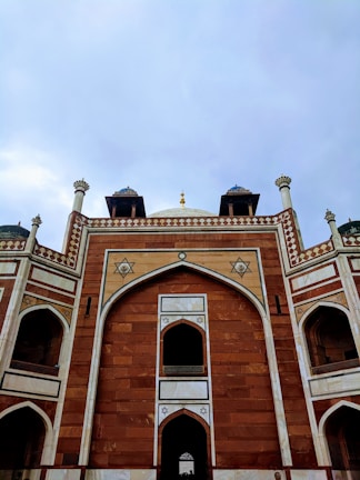 An architectural view of a historical monument featuring intricate geometric patterns and designs. The structure is composed of red sandstone with white marble accents and arches, showcasing traditional craftsmanship. The sky forms a muted backdrop, enhancing the monument's grandeur.