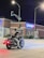 A motorcyclist wearing a black vest with colorful patches is stopped at a nighttime intersection in front of a Chase bank. The scene is illuminated by streetlights and traffic signals, and the motorbike has red lights turned on. The bank building is made of brick and has large windows, with a blue neon sign displaying the word 'CHASE'. There are some plants in front of the bank, and the street signs indicate the intersection of Centralia and 59th Ave.