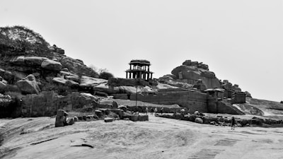 An ancient stone structure stands amidst a rocky terrain, with ruins and large boulders scattered around. Sparse vegetation and a few small trees are visible on the landscape. The architecture has intricate carvings, characteristic of historic temples or monuments.