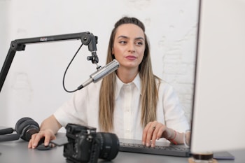 A woman is sitting at a desk, working on a computer while talking into a microphone attached to a boom arm. She is dressed in a white blouse and is focused on her task. A camera and a pair of headphones are also placed on the desk.