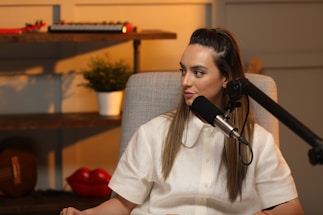 A woman is seated and speaking into a microphone in a cozy room. She is wearing a white shirt and has long hair. Behind her, there are shelves with decorative items, including a small potted plant, a keyboard instrument, and red lips sculpture.