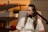 A woman is seated and speaking into a microphone in a cozy room. She is wearing a white shirt and has long hair. Behind her, there are shelves with decorative items, including a small potted plant, a keyboard instrument, and red lips sculpture.