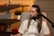 A woman is seated and speaking into a microphone in a cozy room. She is wearing a white shirt and has long hair. Behind her, there are shelves with decorative items, including a small potted plant, a keyboard instrument, and red lips sculpture.