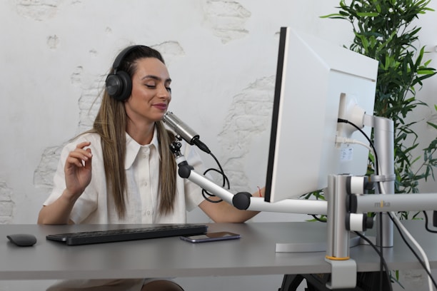 A woman with long hair is sitting at a desk wearing headphones and a white shirt, speaking into a microphone. She appears to be working at a computer with a monitor and a keyboard in front of her. The setting includes a potted plant and walls with a textured, white-washed appearance.