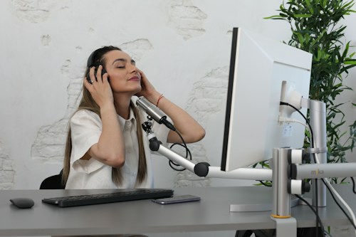A woman wearing headphones sits at a desk in front of a computer monitor. She appears relaxed and is possibly listening to music or a recording. The setting includes a microphone, a keyboard, a mouse, and visible greenery in the background, adding a touch of nature to the workspace.