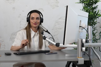 A woman with long hair is wearing headphones and speaking into a microphone at a desk. She is looking at a computer monitor and appears engaged and focused. The room has a minimalist decor with a potted plant and a textured wall.
