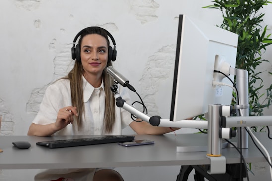 A woman with long hair is wearing headphones and speaking into a microphone at a desk. She is looking at a computer monitor and appears engaged and focused. The room has a minimalist decor with a potted plant and a textured wall.