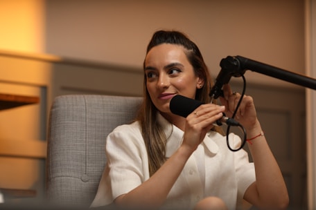 A person is sitting in a comfortable chair, speaking into a microphone. The setting appears to be a studio or recording space with soft lighting and neutral-colored walls. The person is wearing a light-colored blouse and has a relaxed expression.