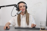 A person is seated at a desk with a microphone setup, wearing headphones and a white button-up shirt, appearing to be involved in a recording or podcasting session. The desk has a keyboard, mouse, and a glass with a beverage.