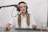 A person is seated at a desk with a microphone setup, wearing headphones and a white button-up shirt, appearing to be involved in a recording or podcasting session. The desk has a keyboard, mouse, and a glass with a beverage.