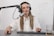 A person is seated at a desk with a microphone setup, wearing headphones and a white button-up shirt, appearing to be involved in a recording or podcasting session. The desk has a keyboard, mouse, and a glass with a beverage.