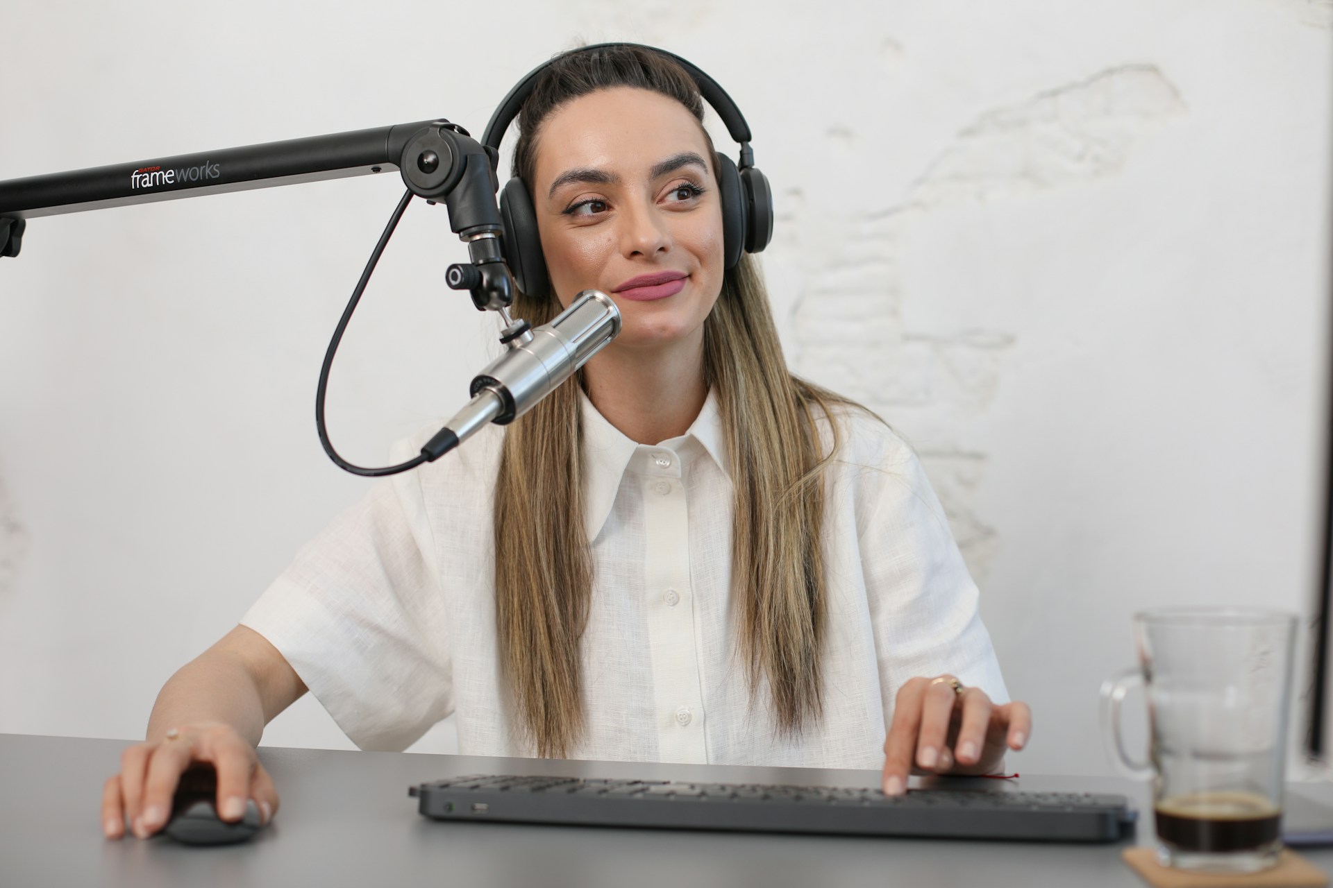 A person is seated at a desk with a microphone setup, wearing headphones and a white button-up shirt, appearing to be involved in a recording or podcasting session. The desk has a keyboard, mouse, and a glass with a beverage.
