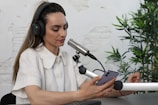 A person wearing headphones and a white shirt is engaged in a podcast or recording session. They are speaking into a professional microphone while holding a smartphone in one hand. The background includes a white brick wall and green plants, creating a relaxed atmosphere.