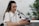 A person wearing headphones and a white shirt is engaged in a podcast or recording session. They are speaking into a professional microphone while holding a smartphone in one hand. The background includes a white brick wall and green plants, creating a relaxed atmosphere.