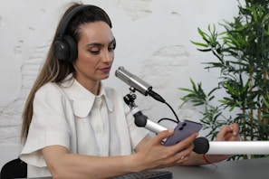 A person wearing headphones and a white shirt is engaged in a podcast or recording session. They are speaking into a professional microphone while holding a smartphone in one hand. The background includes a white brick wall and green plants, creating a relaxed atmosphere.