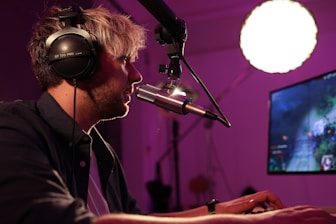A person is sitting at a desk wearing headphones and speaking into a microphone, likely streaming or recording content. The room is dimly lit with a prominent round light source. A computer monitor displays a video game.