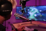 A person wearing headphones is seated in front of a computer screen, engaged in a video game. A microphone is positioned prominently in front of them, suggesting a streaming or recording setup. The background is illuminated with soft purple lighting, creating a cozy and immersive atmosphere.