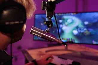 A person wearing headphones is seated in front of a computer screen, engaged in a video game. A microphone is positioned prominently in front of them, suggesting a streaming or recording setup. The background is illuminated with soft purple lighting, creating a cozy and immersive atmosphere.
