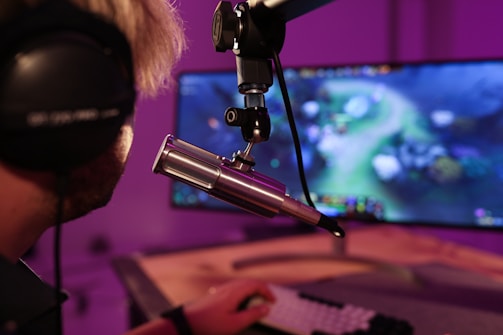 A person wearing headphones is seated in front of a computer screen, engaged in a video game. A microphone is positioned prominently in front of them, suggesting a streaming or recording setup. The background is illuminated with soft purple lighting, creating a cozy and immersive atmosphere.