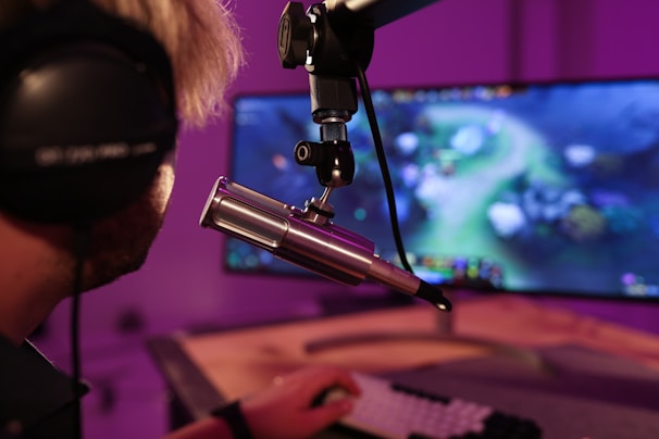 A person wearing headphones is seated in front of a computer screen, engaged in a video game. A microphone is positioned prominently in front of them, suggesting a streaming or recording setup. The background is illuminated with soft purple lighting, creating a cozy and immersive atmosphere.