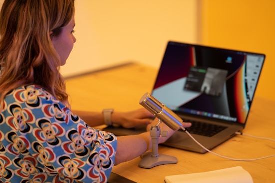 A woman with long hair sits at a wooden table using a laptop. Her focus is on the screen. A silver microphone on a stand is positioned near her, and a notebook or journal sits nearby. The atmosphere appears to be warm with soft lighting.