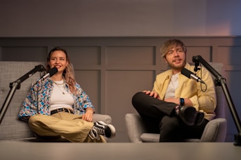 Two people are seated in comfortable chairs inside a studio setting, with microphones positioned in front of them, suggesting they are hosting or participating in a podcast or interview. One person is wearing a colorful patterned shirt and khaki pants, while the other is dressed in a light yellow jacket and black trousers. The background consists of a wall with a panel design, and there is soft lighting adding warmth to the ambiance.