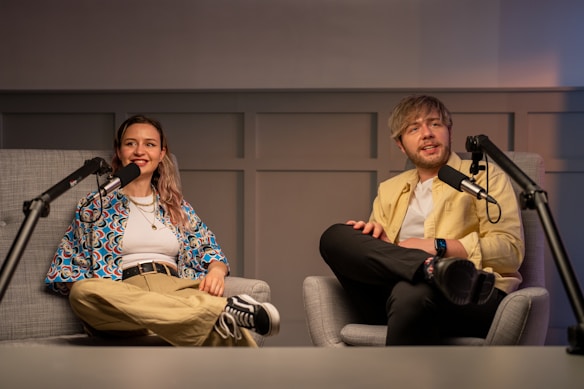 Two people are seated in comfortable chairs inside a studio setting, with microphones positioned in front of them, suggesting they are hosting or participating in a podcast or interview. One person is wearing a colorful patterned shirt and khaki pants, while the other is dressed in a light yellow jacket and black trousers. The background consists of a wall with a panel design, and there is soft lighting adding warmth to the ambiance.