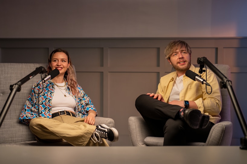 Two people are seated in comfortable chairs inside a studio setting, with microphones positioned in front of them, suggesting they are hosting or participating in a podcast or interview. One person is wearing a colorful patterned shirt and khaki pants, while the other is dressed in a light yellow jacket and black trousers. The background consists of a wall with a panel design, and there is soft lighting adding warmth to the ambiance.