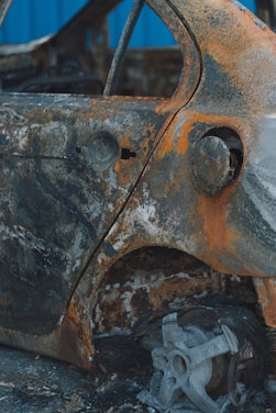 A heavily rusted and damaged car with significant corrosion on its surface. The metal is oxidized with orange and brown hues, exposing the frame and wheel well. The wheel is missing, revealing the car's structural elements.