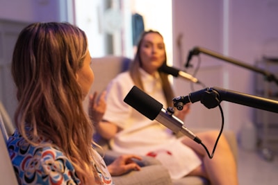Two women are seated in a studio setting, engaged in conversation with professional microphones positioned between them. One woman is in focus with long hair and a patterned shirt while the other is slightly blurred in the background.