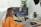 A person with long hair and an orange shirt works at a desk with audio-visual editing equipment. They are speaking into a microphone that is attached to an adjustable arm. A large monitor displays video editing software, and wireless headphones are placed on the desk. In the background, there are potted plants and a brick wall.