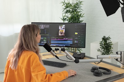 A person with long hair and an orange shirt works at a desk with audio-visual editing equipment. They are speaking into a microphone that is attached to an adjustable arm. A large monitor displays video editing software, and wireless headphones are placed on the desk. In the background, there are potted plants and a brick wall.