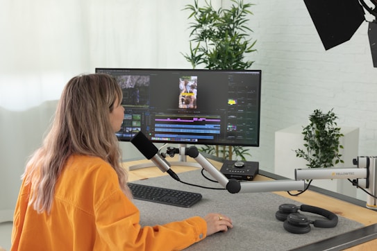A person with long hair and an orange shirt works at a desk with audio-visual editing equipment. They are speaking into a microphone that is attached to an adjustable arm. A large monitor displays video editing software, and wireless headphones are placed on the desk. In the background, there are potted plants and a brick wall.