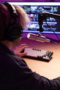 A person wearing headphones sits in front of a computer monitor with a professional microphone mounted on a boom arm. The screen displays several video thumbnails, and the person is typing on a mechanical keyboard. The setting has a warm, ambient purple and pink lighting.