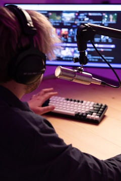 A person wearing headphones sits in front of a computer monitor with a professional microphone mounted on a boom arm. The screen displays several video thumbnails, and the person is typing on a mechanical keyboard. The setting has a warm, ambient purple and pink lighting.