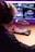 A person wearing headphones sits in front of a computer monitor with a professional microphone mounted on a boom arm. The screen displays several video thumbnails, and the person is typing on a mechanical keyboard. The setting has a warm, ambient purple and pink lighting.