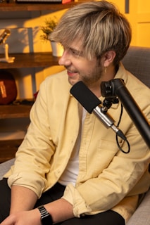 A person with light-colored hair is seated in a cozy room, speaking into a microphone. They are wearing a yellow shirt and a smartwatch. There is a shelf in the background with various items including a guitar, a potted plant, and other decorations. The lighting is warm and inviting, adding to the overall relaxed atmosphere.