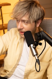 A person with light brown hair and a beard is sitting in a chair, engaged in speaking or singing into a microphone attached to a stand. They are wearing a yellow shirt over a white t-shirt and an earring is visible on the left ear. The background has a warm, cozy ambiance with wooden shelves visible.