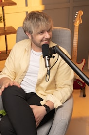 A person with blonde hair is seated in a light gray armchair, smiling, holding a microphone, and dressed in a yellow shirt over a white t-shirt. There is an electric guitar leaning against the wall and a wooden shelf with a potted plant in the background.