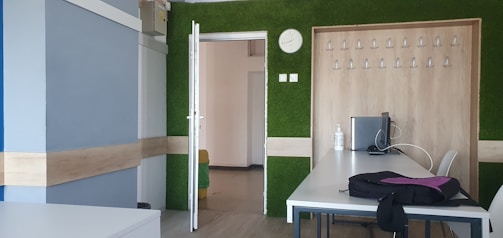 A modern office room featuring minimalist design with a mix of green, wood, and blue elements. The room includes a closed laptop, a clock showing approximately 10:10, a row of coat hooks on a feature wall with a green texture, and a desk with a bottle, a computer monitor, and a chair. There is an open door leading to a hallway.