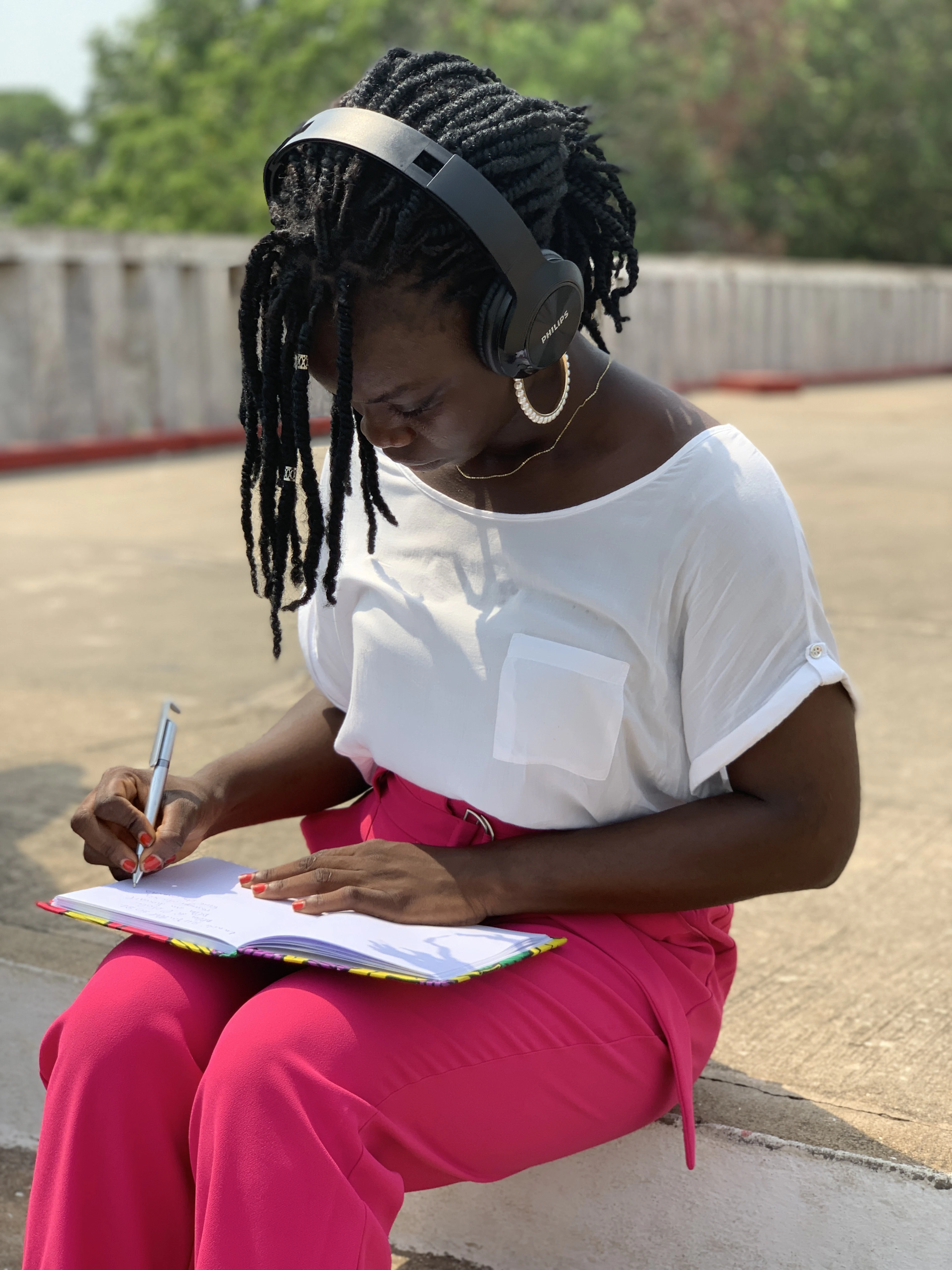 A woman in a white top and pink trousers sitting outdoors writing in a notebook