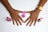 A pair of hands with freshly painted nails in a pinkish-purple color, with three bottles of nail polish placed beside them. One hand wears a gold wristwatch.