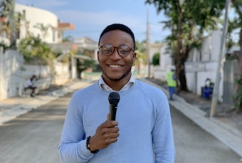 A person is standing in the middle of a street holding a microphone. The individual is wearing glasses and a light blue sweater, smiling warmly. The background shows a residential neighborhood with trees, a white fence, and two people in the distance, one sitting and the other walking.