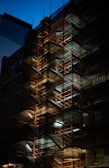 A multi-level scaffolding structure is illuminated against the backdrop of a darkened urban environment. The metal framework is intricately woven with horizontal and vertical lines, creating a geometric pattern. Lights are positioned at various levels, casting shadows and highlighting the network of stairs and platforms. The contrast between the warm tones of the lit scaffolding and the cool, dark tones of the surrounding buildings and sky creates a dramatic visual effect.